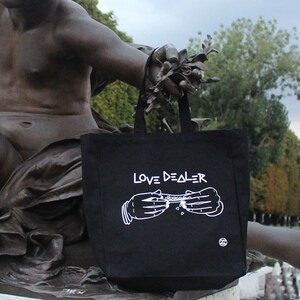 May include: Black canvas tote bag with white text that reads "LOVE DEALER" and a graphic of two hands rolling a joint.