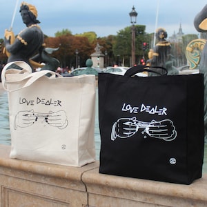 May include: Two tote bags with the text "LOVE DEALER" printed on them. One bag is black with white text and the other is white with black text. Both bags feature a graphic of two hands rolling a joint.