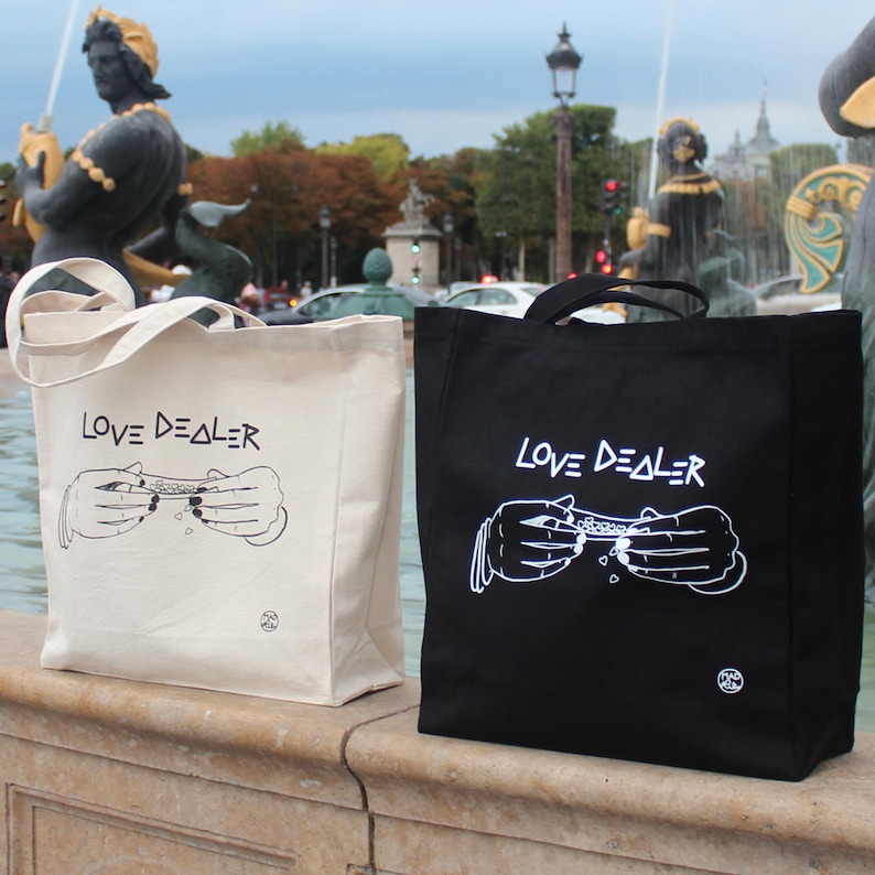 May include: Two tote bags with the text "LOVE DEALER" printed on them. One bag is black with white text and the other is white with black text. Both bags feature a graphic of two hands rolling a joint.