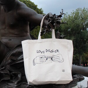 May include: A white canvas tote bag with black text that reads "LOVE DEALER". The bag features a black and white illustration of two hands rolling a joint.