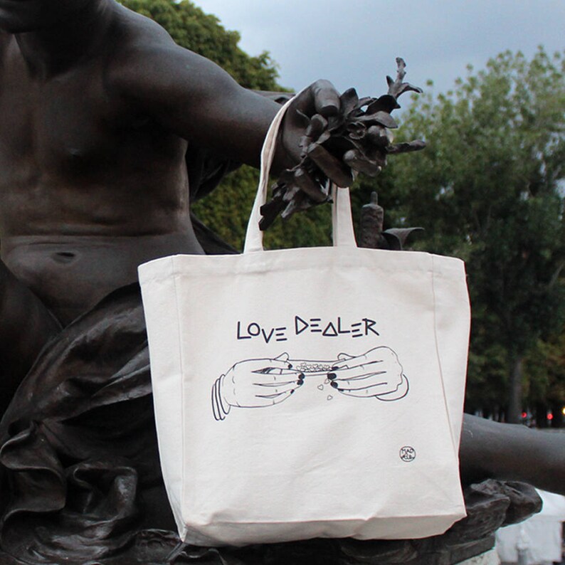 May include: A white canvas tote bag with black text that reads "LOVE DEALER". The bag features a black and white illustration of two hands rolling a joint.