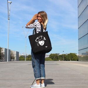 May include: A woman wearing blue jeans and a black and white patterned shirt is carrying a black tote bag with a white illustration of a woman's face on it. The bag has a white text that says "#88".