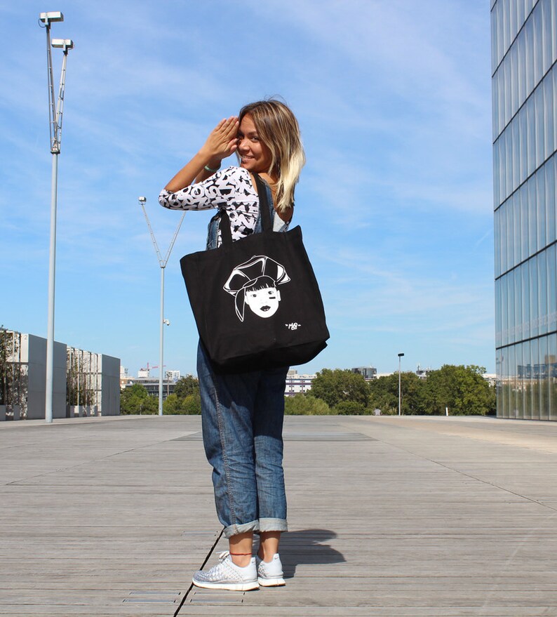 May include: A woman wearing blue jeans and a black and white patterned shirt is carrying a black tote bag with a white illustration of a woman's face on it. The bag has a white text that says "#88".