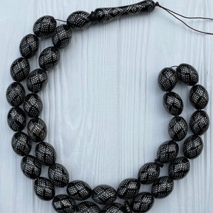 May include: A black prayer beads necklace with silver accents. The beads are oval shaped and have a geometric pattern. The necklace is strung on a brown cord.