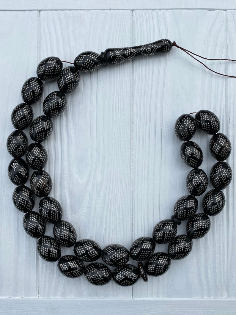 May include: A black prayer beads necklace with silver accents. The beads are oval shaped and have a geometric pattern. The necklace is strung on a brown cord.