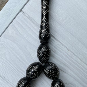 May include: A black prayer beads necklace with silver accents. The beads are oval shaped and have a repeating pattern of small silver dots. The necklace has a tassel at the end.
