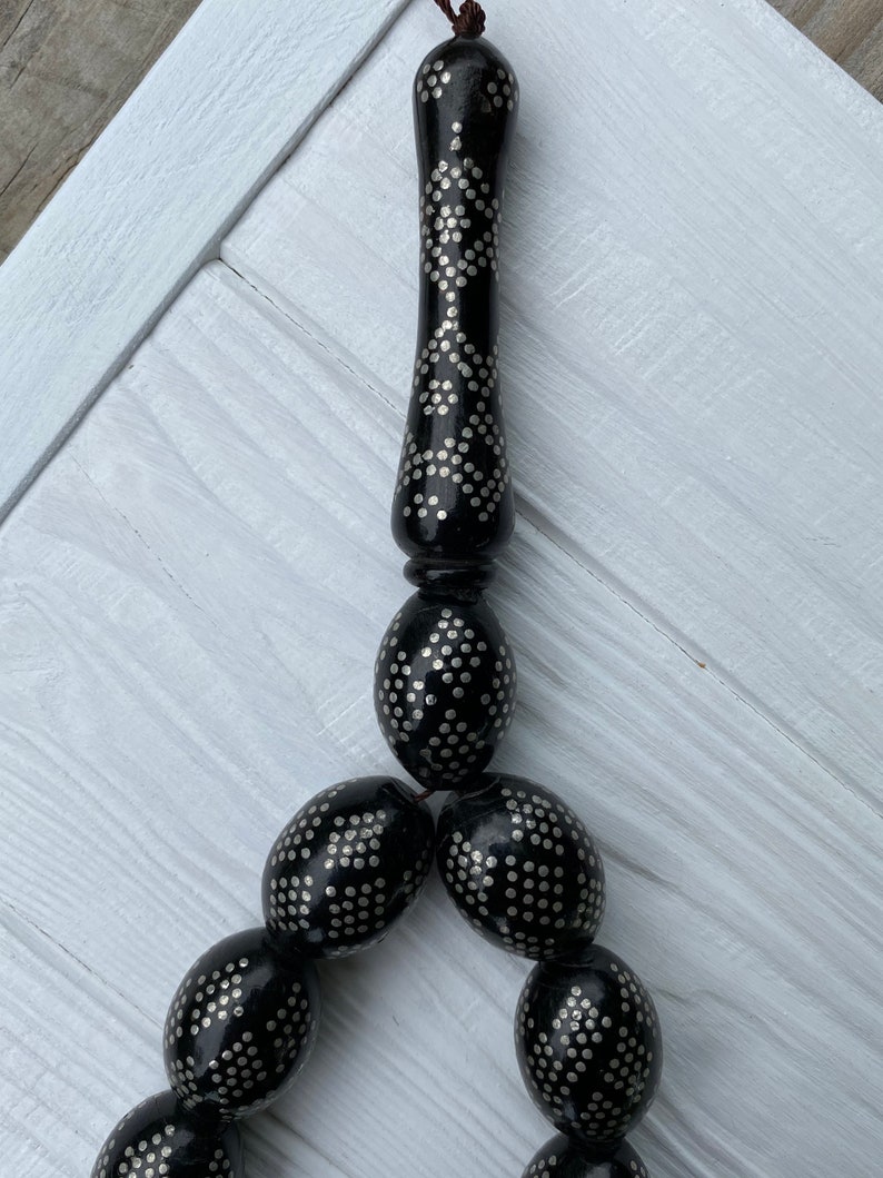May include: A black prayer beads necklace with silver accents. The beads are oval shaped and have a repeating pattern of small silver dots. The necklace has a tassel at the end.