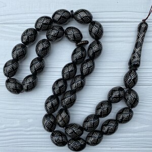 May include: A black prayer beads necklace with silver accents. The beads are oval shaped and have a geometric pattern. The necklace is strung on a black cord.