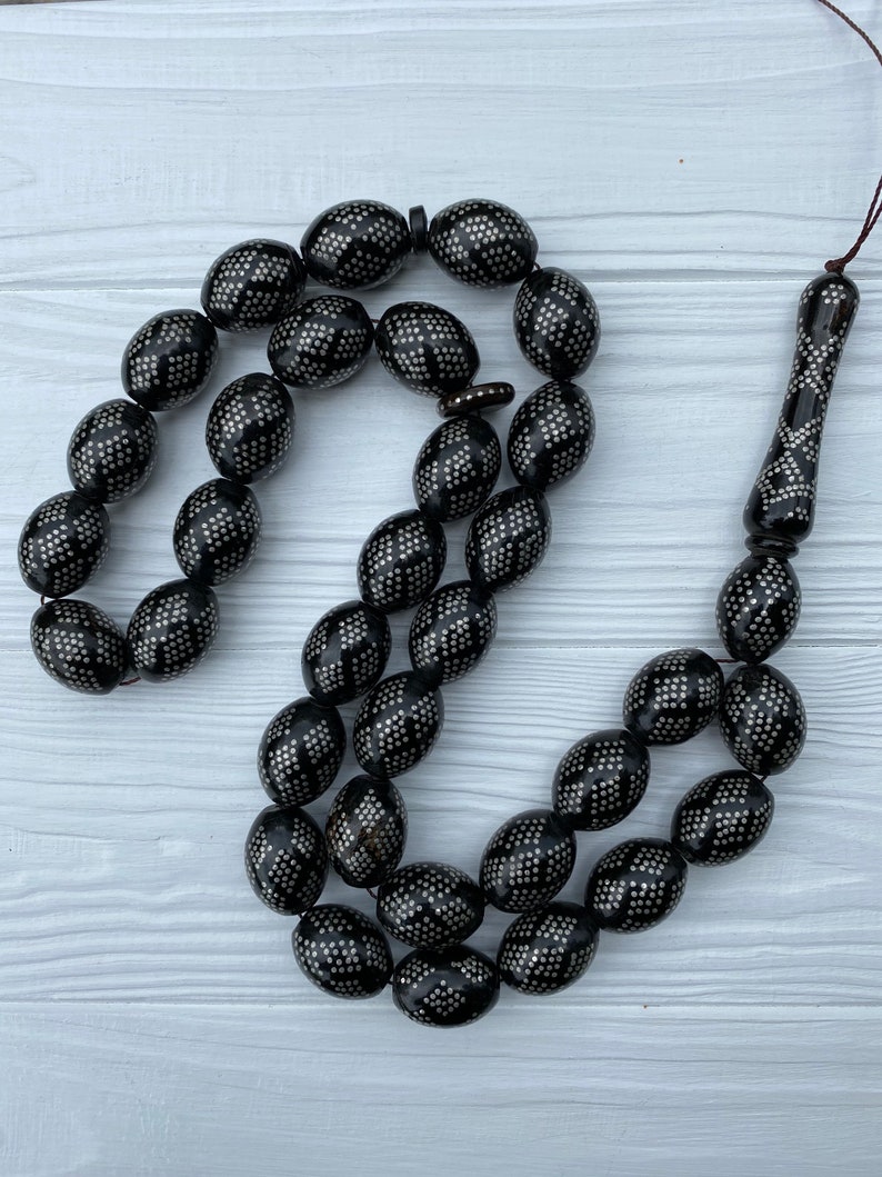 May include: A black prayer beads necklace with silver accents. The beads are oval shaped and have a geometric pattern. The necklace is strung on a black cord.