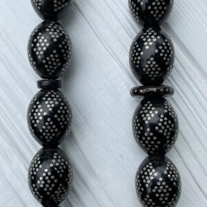 May include: A black prayer bead necklace with silver accents. The beads are oval shaped and have a repeating pattern of small silver dots.