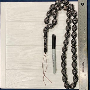 May include: A black prayer beads necklace with silver accents and a tassel. The beads are oval shaped and have a geometric pattern. The necklace is laid out on a white wooden surface with a black marker and a ruler.