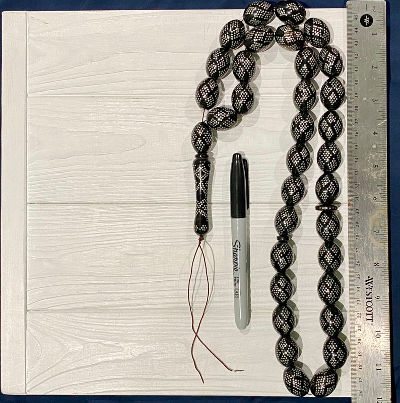 May include: A black prayer beads necklace with silver accents and a tassel. The beads are oval shaped and have a geometric pattern. The necklace is laid out on a white wooden surface with a black marker and a ruler.