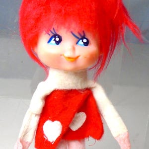May include: A vintage doll with a red felt dress and matching boots. The doll has a white collar, red hair, and a smiling face with blue eyes. The dress features two white heart-shaped accents.