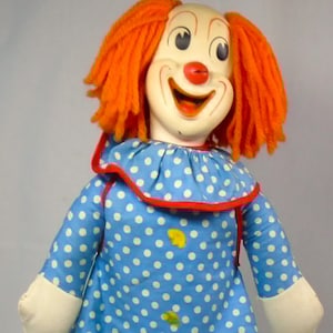 May include: A clown doll with bright orange yarn hair, a painted face featuring a red nose, and a wide smile. The doll wears a blue dress and matching pants with white polka dots, a ruffled collar with red trim, and solid yellow shoes.