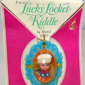 May include: Vintage Lucky Locket Kiddle by Mattel. The locket features a small doll inside an oval frame with a blue border. The packaging is pink and white with the text "Wear me and be lucky!"