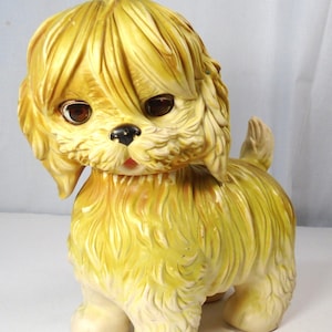 May include: A vintage ceramic dog figurine in shades of yellow and cream. The dog has large brown eyes, a black nose, and a pink mouth. Its fur is textured, and it stands on four cream-colored paws. The figurine has a retro aesthetic.