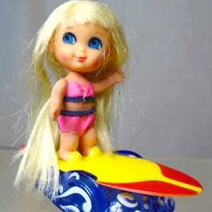 May include: A vintage doll with blonde hair and blue eyes, wearing a pink and blue swimsuit, stands on a colorful surfboard. The surfboard is yellow, red, and blue with white decorative swirls. A retro toy.
