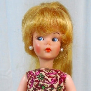 May include: A vintage doll with blonde hair, blue eyes, and rosy cheeks. She wears a patterned crop top with pink, green, and brown designs, and a pair of hot pink shorts. The doll has pearl earrings and is standing.