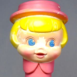 May include: A molded plastic figurine of a character with bright yellow hair, wearing a pink hat, a pink collared dress with long sleeves, and pink shoes. The character has blue eyes, rosy cheeks, and a small mouth, with hands clasped together.