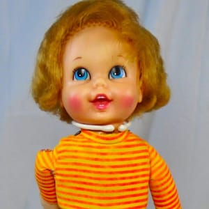 May include: A vintage doll with a cheerful expression, blue eyes, and rosy cheeks. The doll wears a yellow and orange striped shirt, a yellow skirt, orange leggings, and white boots. The doll is approximately 12 inches tall.
