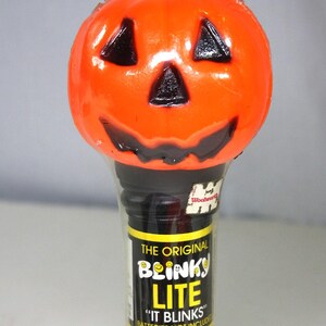 May include: An orange pumpkin-shaped Halloween light with black facial features. The black base is wrapped in plastic and has a yellow and black label that reads "The Original Blinky Lite".