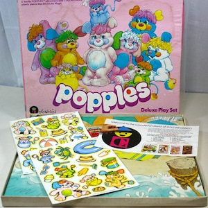 May include: A vintage Popples Deluxe Play Set box with colorful cartoon characters. The box is pink with the word "Popples" in large, white letters. Stickers and a play scene are visible inside the box.