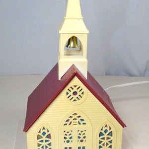 May include: A miniature church model with a steeple, bell tower, and a burgundy roof. The church has arched windows with colorful stained-glass-style inserts. The building is painted in a light yellow color and sits on a beige base.