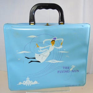 May include: A vintage light blue suitcase featuring a graphic of a nun in a white habit, flying among clouds. The suitcase has a black handle and the text "THE FLYING NUN". A church is depicted at the bottom.