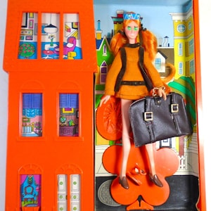 May include: Vintage doll in an orange plastic house-shaped case. The doll has red hair, green eyes, and wears an orange outfit with a black belt and a brown purse. The case has windows with interior scenes and a cityscape backdrop.