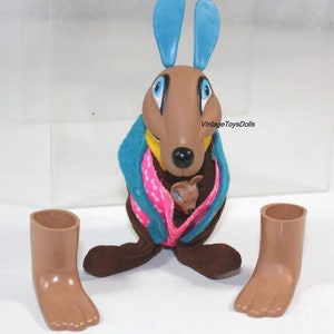 May include: A brown plush kangaroo toy with blue ears, holding a smaller brown plush kangaroo joey. The kangaroo is wearing a teal blue and pink patterned scarf. The toy is missing its feet, which are shown separately in the image.