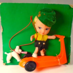 May include: A small doll with blonde hair wearing a green hat, yellow top, and black shorts, standing on an orange plastic scooter. A white dog figure is attached by a thin brown cord. The set is mounted on a square green cardboard backing.