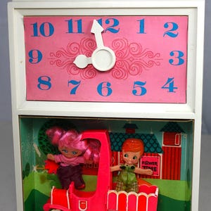 May include: A vintage toy clock with a pink face and blue numbers. The clock has a movable hand and a small window below it that shows a red toy truck with two dolls inside. The dolls are a girl with pink hair and a boy with red hair. The truck has a sign that says "Flower Time".