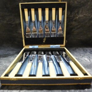 May include: A vintage silverware set in a wooden box. The set includes forks and knives with cream-colored handles and silver blades and tines. The box is lined with blue satin and black velvet.