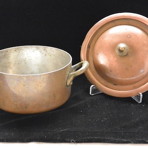 May include: A round copper pot with two side handles and a matching circular lid. The pot has a metallic interior finish, while the exterior and the lid display a warm, weathered copper patina with visible signs of age and use throughout.
