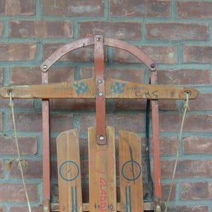 May include: Vintage wooden sled with red metal frame and rope handles. The sled has a wooden seat with a "Gladding" logo and "C.M.S." written on the front.