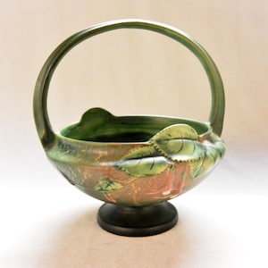 May include: A ceramic basket with a handle, featuring a green and brown color scheme. The basket is adorned with leaf-shaped details and rests on a dark base. The handle arches gracefully above the bowl.