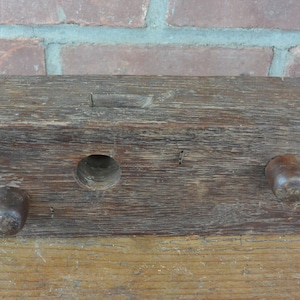 May include: A weathered, rectangular wooden object with a dark brown finish. It features a central circular hole, a small rectangular slot, and two rounded knobs on either end. The wood grain is visible, and the piece appears aged.
