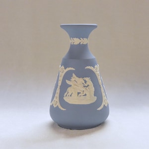 May include: A light blue ceramic vase with white relief detailing. The vase features a classical scene with figures and decorative floral accents. The vase has a flared top and a rounded base.