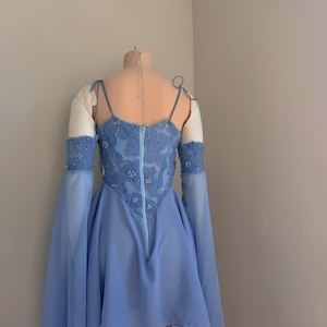 Puede incluir: Un vestido azul claro con un corpi&ntilde;o ajustado y una falda fluida, exhibido en un maniqu&iacute;. El corpi&ntilde;o est&aacute; adornado con intrincados abalorios y presenta una cremallera en la espalda. Mangas largas y transparentes caen de los hombros.
