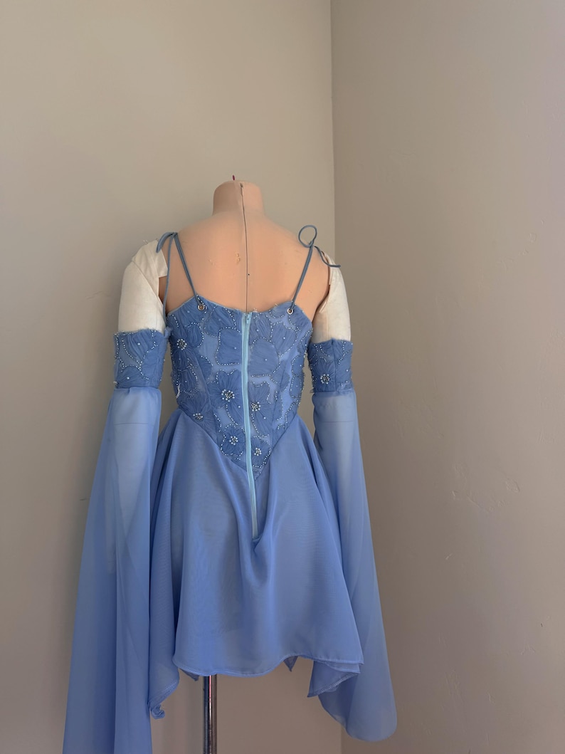 Puede incluir: Un vestido azul claro con un corpi&ntilde;o ajustado y una falda fluida, exhibido en un maniqu&iacute;. El corpi&ntilde;o est&aacute; adornado con intrincados abalorios y presenta una cremallera en la espalda. Mangas largas y transparentes caen de los hombros.