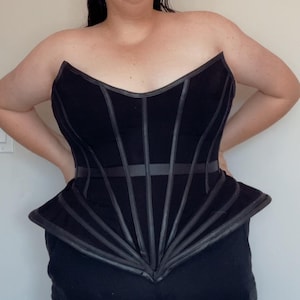 Puede incluir: Un cors&eacute; negro sin tirantes con detalles verticales y horizontales de aspecto cuero. El cors&eacute; tiene un dise&ntilde;o estructurado y ajustado con una parte inferior estilo peplum. La prenda est&aacute; hecha de una tela oscura y presenta un escote en V.