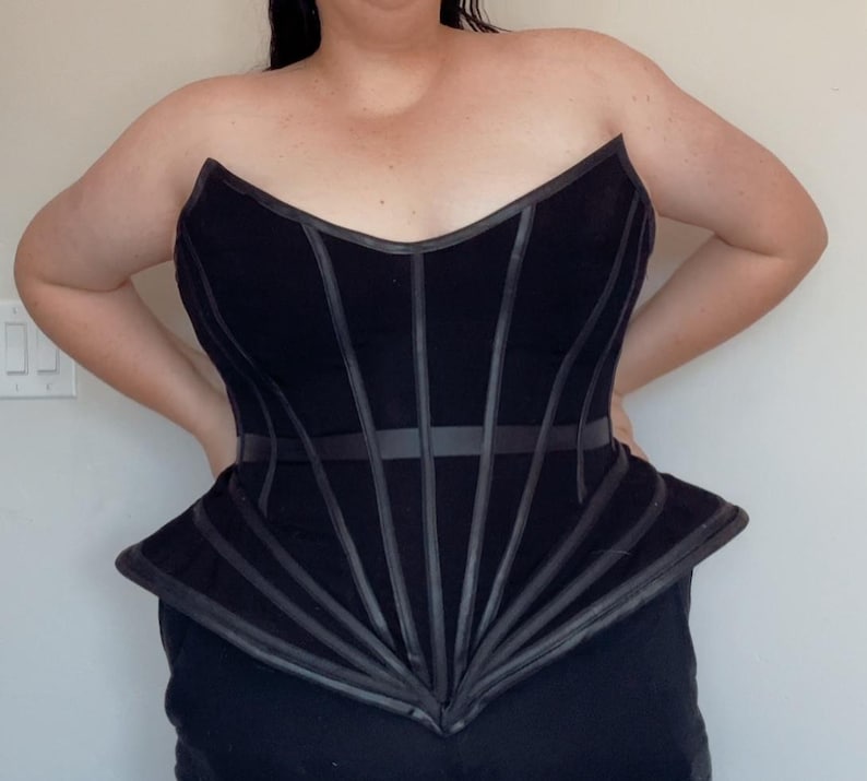 Puede incluir: Un cors&eacute; negro sin tirantes con detalles verticales y horizontales de aspecto cuero. El cors&eacute; tiene un dise&ntilde;o estructurado y ajustado con una parte inferior estilo peplum. La prenda est&aacute; hecha de una tela oscura y presenta un escote en V.