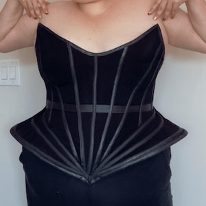 Puede incluir: Cors&eacute; negro con un dise&ntilde;o estructurado. El corpi&ntilde;o presenta l&iacute;neas verticales y horizontales, con un peplum acampanado. El cors&eacute; tiene un escote coraz&oacute;n y est&aacute; hecho de una tela oscura con detalles de cuero.