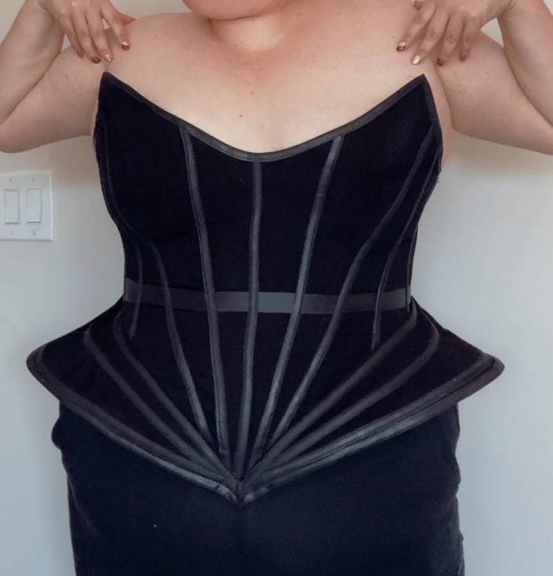 Puede incluir: Cors&eacute; negro con un dise&ntilde;o estructurado. El corpi&ntilde;o presenta l&iacute;neas verticales y horizontales, con un peplum acampanado. El cors&eacute; tiene un escote coraz&oacute;n y est&aacute; hecho de una tela oscura con detalles de cuero.