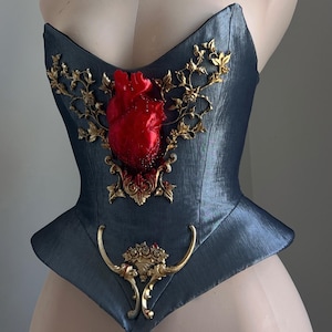 Puede incluir: Un cors&eacute; azul verdoso oscuro con un coraz&oacute;n rojo esculpido y adornos dorados. El coraz&oacute;n es el punto focal, rodeado de filigranas doradas ornamentadas. El cors&eacute; tiene una forma estructurada y ajustada.