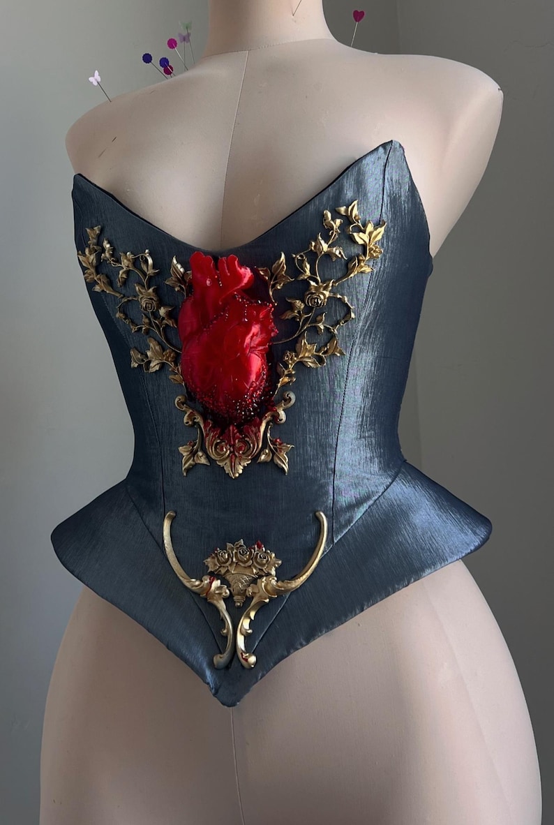 Puede incluir: Un cors&eacute; azul verdoso oscuro con un coraz&oacute;n rojo esculpido y adornos dorados. El coraz&oacute;n es el punto focal, rodeado de filigranas doradas ornamentadas. El cors&eacute; tiene una forma estructurada y ajustada.