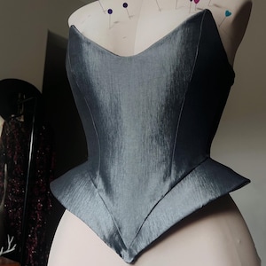 Puede incluir: Un cors&eacute; gris con un dise&ntilde;o estructurado y angular est&aacute; prendido a un maniqu&iacute;. El cors&eacute; tiene un escote en V y una cintura estilo peplum. La tela tiene un brillo sutil y el maniqu&iacute; est&aacute; cubierto de alfileres.