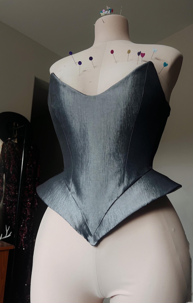 Puede incluir: Un cors&eacute; gris con un dise&ntilde;o estructurado y angular est&aacute; prendido a un maniqu&iacute;. El cors&eacute; tiene un escote en V y una cintura estilo peplum. La tela tiene un brillo sutil y el maniqu&iacute; est&aacute; cubierto de alfileres.