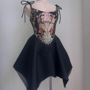 Puede incluir: Vestido negro con corpi&ntilde;o estampado floral y falda acampanada. El corpi&ntilde;o presenta bordados florales dorados, rosas y rojos sobre un fondo negro. El vestido tiene tirantes anudados y se exhibe en un maniqu&iacute;.