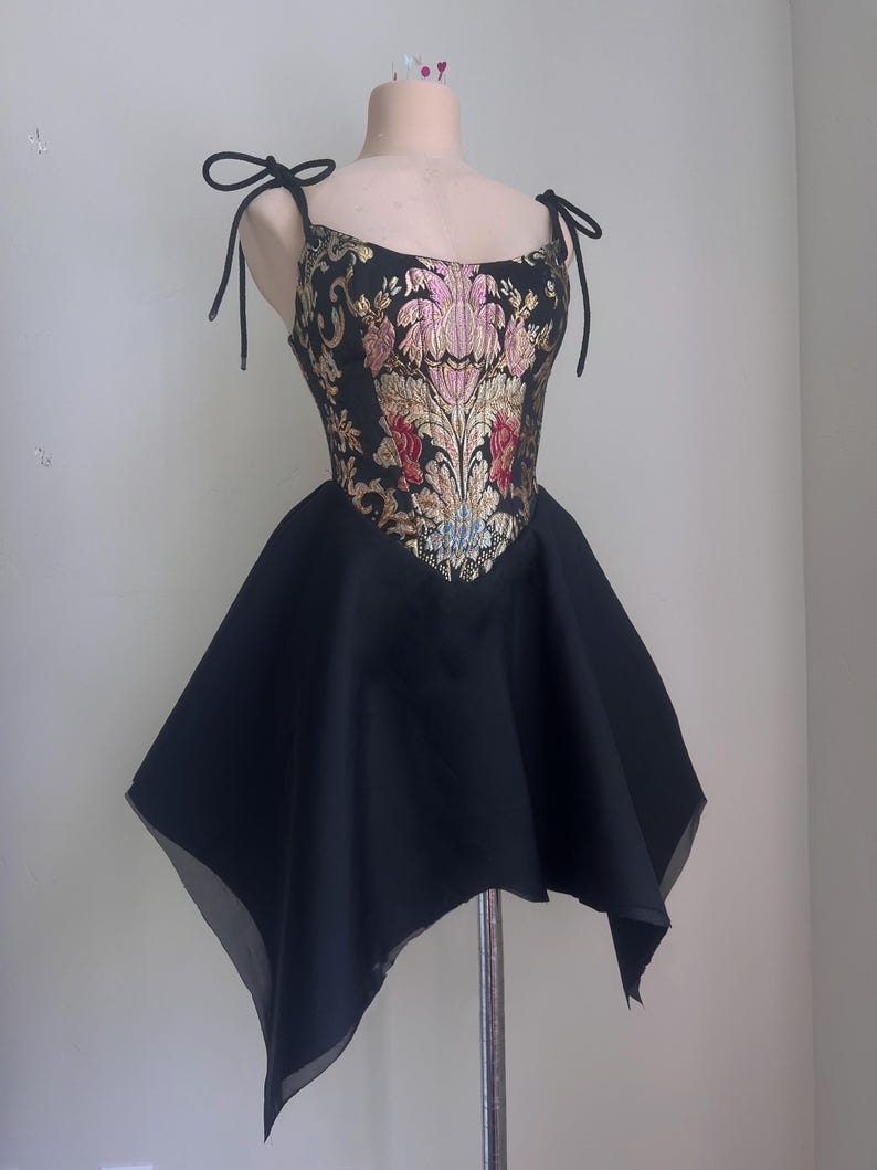 Puede incluir: Vestido negro con corpi&ntilde;o estampado floral y falda acampanada. El corpi&ntilde;o presenta bordados florales dorados, rosas y rojos sobre un fondo negro. El vestido tiene tirantes anudados y se exhibe en un maniqu&iacute;.
